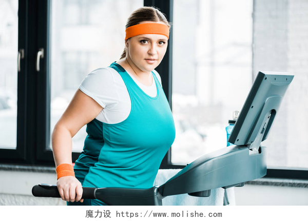 在跑步机上健身的女孩有吸引力的超重女孩在跑步机上锻炼, 而看着相机在健身房